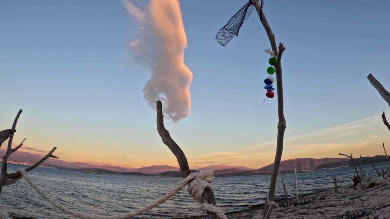Sereno paesaggio costiero con scultura artistica in legno漂流legno con pietre colorate appese e rete da pesca su spiaggia di ciottoli durante un tramonto suggestivo con nuvole sopra il mare calmo e montagne in lontananza