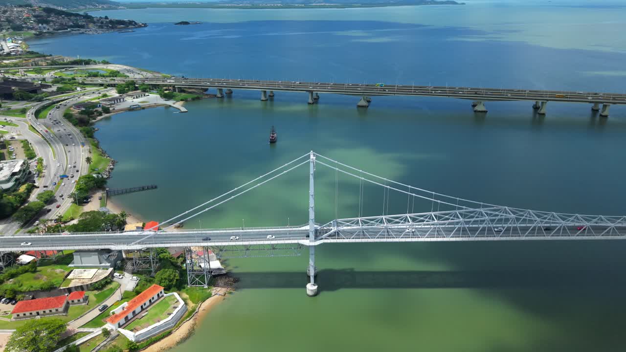 Tomada de drone ascendente da histórica ponte Hercílio Luz. A cena mostra arquitetura urbana, casas costeiras, rodovia e tráfego constante atravessando a água sob uma mistura de sol e nuvens.