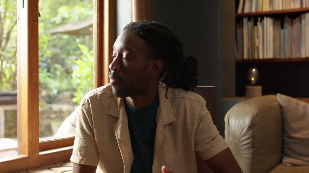 African man in beige shirt on couch holding white mug gazing outside turning toward camera smiling