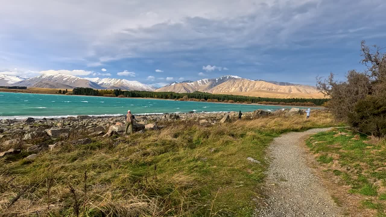 Two people walk slowly along a gravel path beside a turquoise lake, surrounded by grass, thistles, and mountains under cloudy daylight. Camera pans smoothly