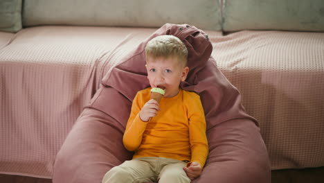 cute little boy relaxing on pink bean bag chair, wearing bright yellow shirt, happily eating chocolate-covered mint ice cream cone, cozy indoor setting with soft background