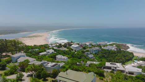 Diapositiva-Y-Toma-Panorámica-De-Casas-Familiares-Y-Residencias-Cerca-De-La-Costa.-Olas-Rodando-Sobre-La-Superficie-Del-Agua-Hasta-La-Costa.-Bahía-Plettenberg,-Sudáfrica