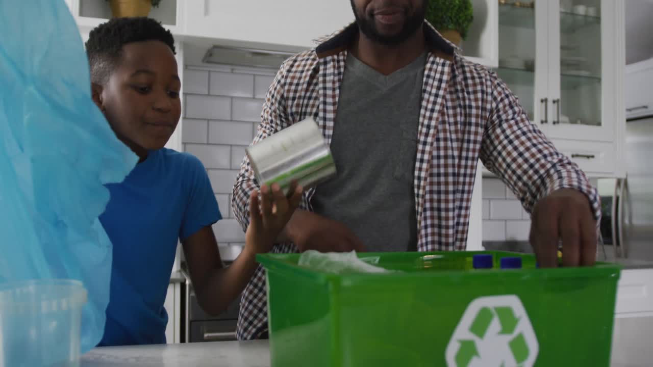 feliz padre y hijo afroamericanos de pie en la cocina clasificando basura en la caja de reciclaje