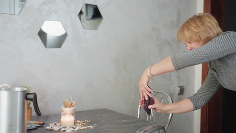 young woman adjusting backward smiling as she takes picture of cozy candlelight decor with phone upside down, surrounded by seashells, decorative sticks, metal jug, small potted plant on gray table
