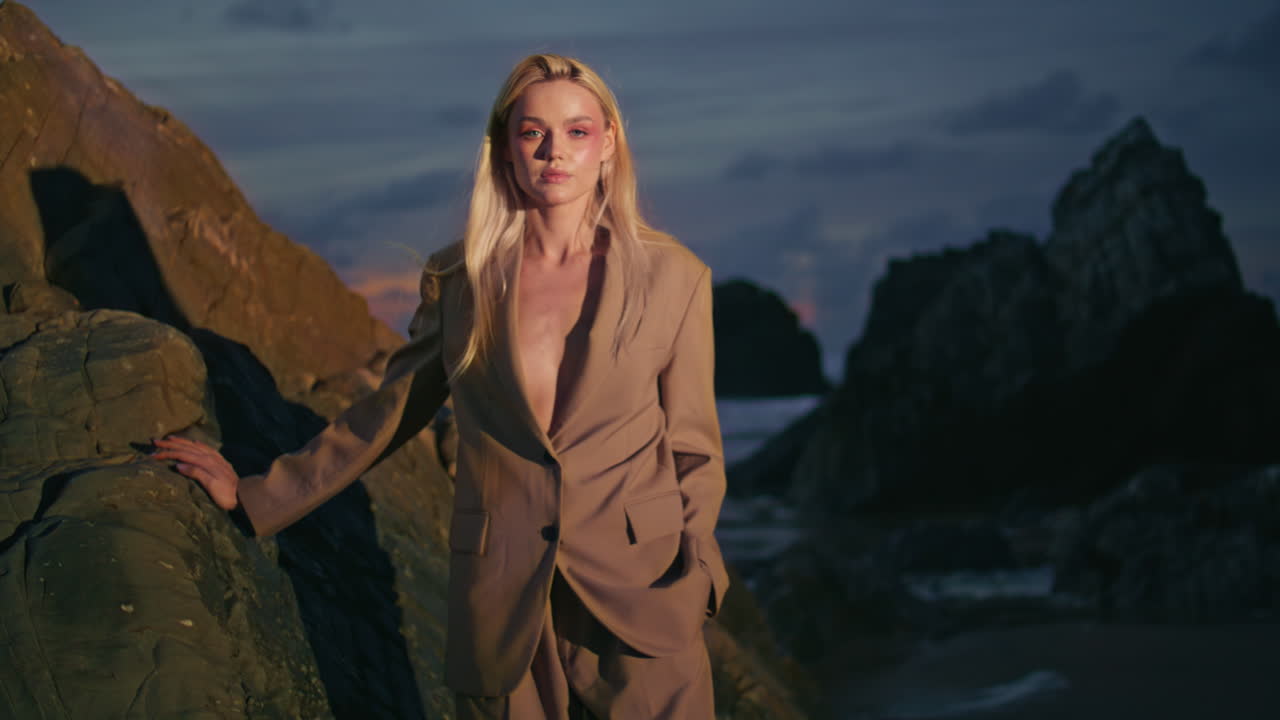 어두운 저녁에 모델링하는 패션 소녀 해안. 섹시하고 자신감있는 금발이 바위 포즈를 취합니다.