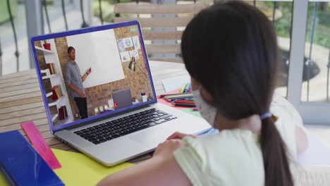 colegiala caucásica con máscara facial usando una computadora portátil en una videollamada con un profesor masculino