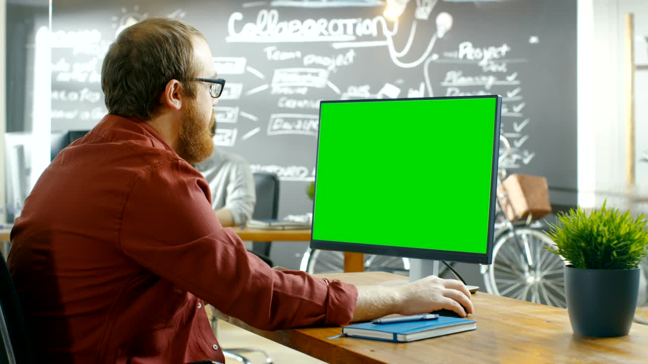 el hombre trabaja en la computadora personal con pantalla de maqueta verde. en el fondo oficina creativa con pared de pizarra.