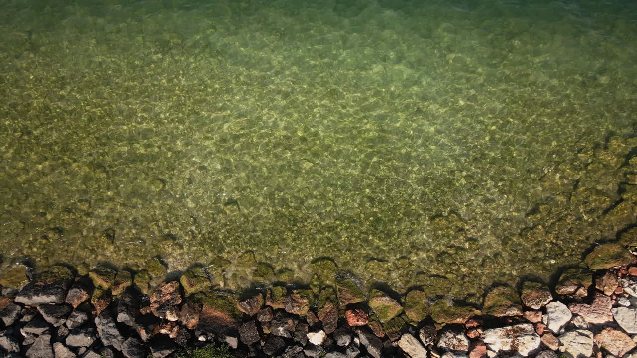 Smooth 4K drone ascent from a close top-down view of Lake Garda rocks, rising to reveal more of the clear water and shoreline as waves wash over the stones