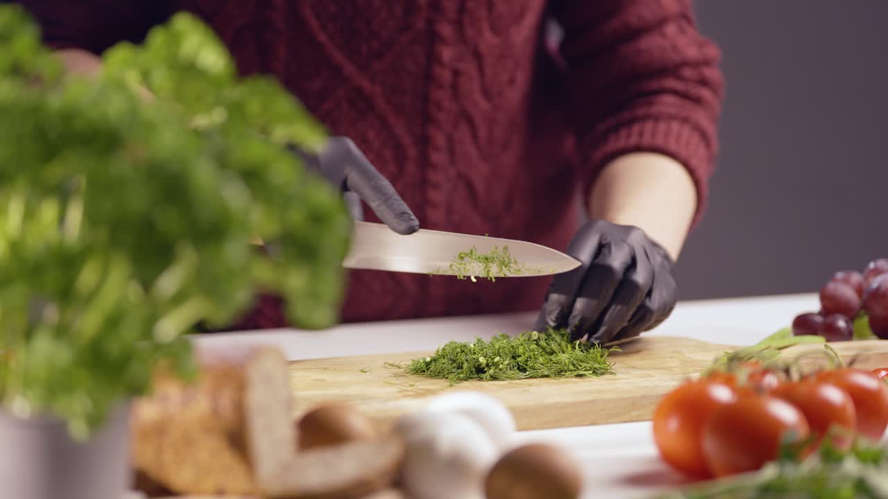 hands wearing black gloves finely chopping fresh dill on a wooden cutting board. A sharp knife slices through the green herb, creating delicate pieces. Fresh ingredients surround the workspace
