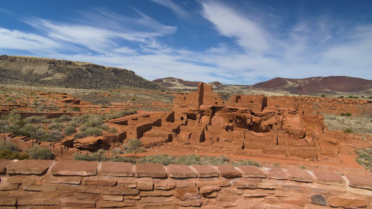 un tiro de gran angular que se eleva desde un muro de piedra, revelando las ruinas más grandes del pueblo rodeadas de matorrales del desierto en el monumento nacional wupatki en arizona