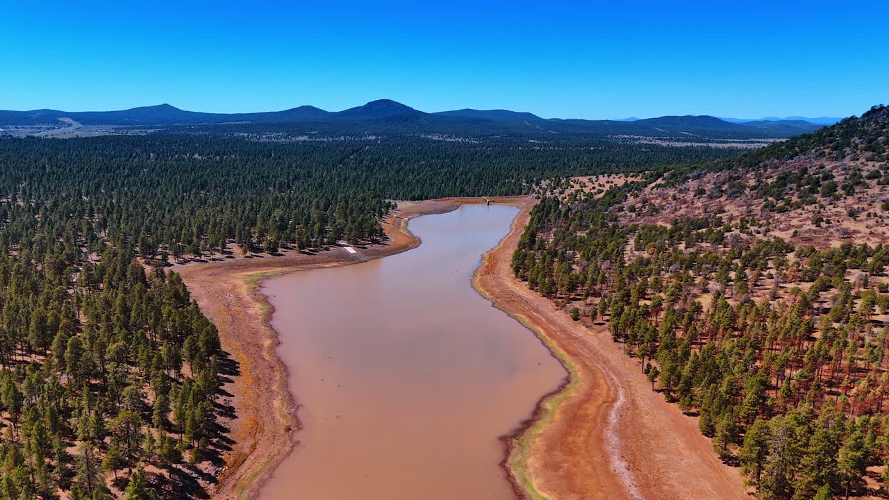 Footage over the shallow river with brown water and banks. Pine trees grow around in the valley and on the mountains. Scenery of Arizona, USA