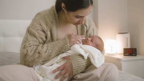 front view of brunette smiling woman sitting on the bed holding her baby and caressing him