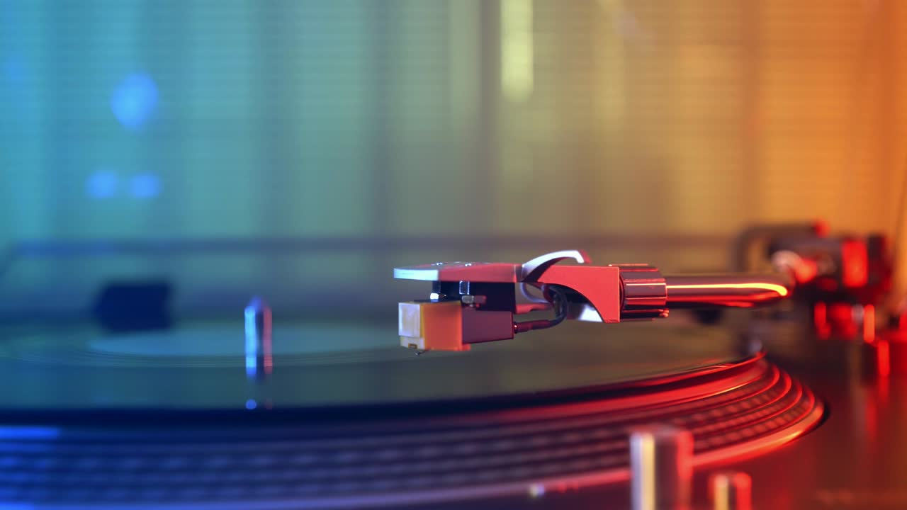 비닐 플레이어 턴테이블: 회전하는 비닐 레코드의 조각, 바늘을 클로즈업으로 가진 스타일러스, 바늘과 비닐 판을 멈추는, 매크로, 전면 뷰, 빨간색과 파란색 빛의 배경에서 흐릿한.