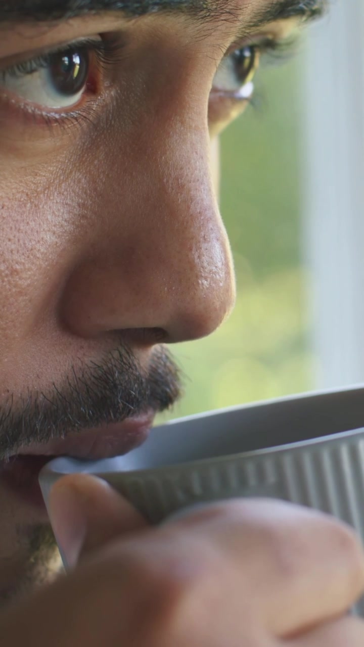 Video verticale: Uomo indiano che sorseggia, stimolato dalla luce del giorno, che alza una tazza a coste e guarda fuori dalla finestra.