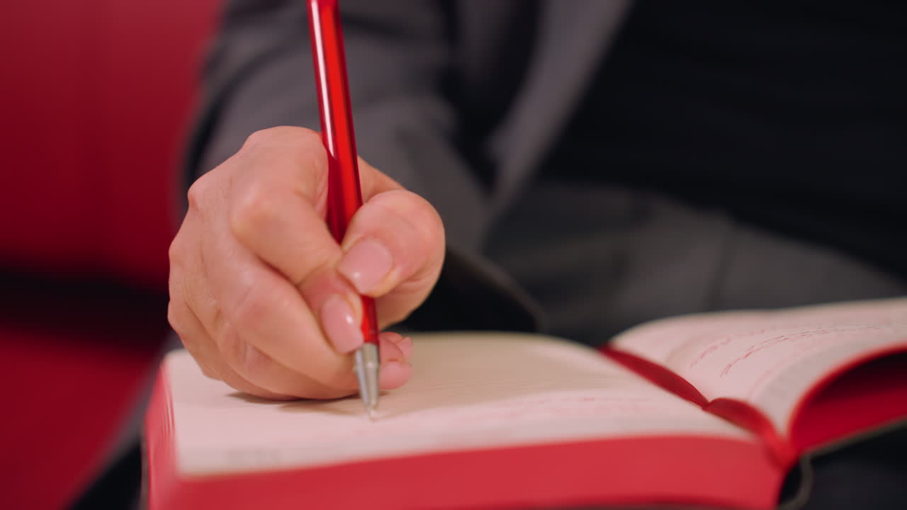 Close-up of hand holding red pen writing in open notebook. Focus on journaling, note-taking, and professional planning. Creative workspace, productive business environment, action in modern office