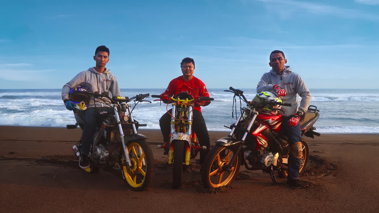 Portrait of three motocross stunt riders sitting on their motorcycles, off-road at the beach with the ocean behind them.