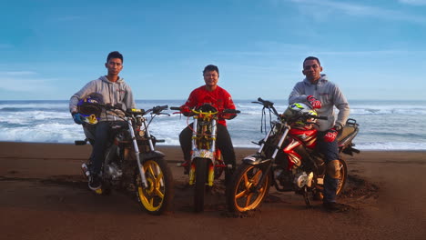 portrait of three motocross stunt riders sitting on their motorcycles, off-road at the beach with the ocean behind them.