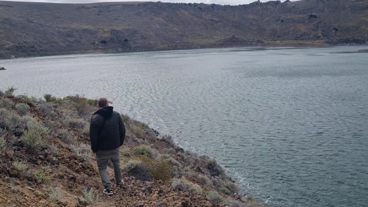 Un escursionista solitario esplora le rive vulcaniche della Laguna Azul vicino a Río Gallegos, Argentina. Cieli tempestosi circondano questo lago vulcanico nel campo vulcanico di Pali Aike, mostrando il paesaggio selvaggio della Patagonia.