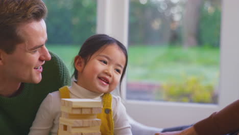 Tochter-Flüstert-Den-Eltern-Ins-Ohr,-Während-Die-Familie-Mit-Zwei-Vätern-Ein-Spiel-Spielt-Und-Holzsteine-Stapelt