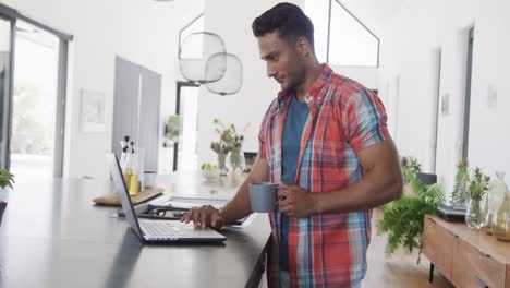 Hombre-Birracial-Tomando-Café-Y-Usando-Una-Computadora-Portátil-En-Un-Apartamento-Moderno-Y-Soleado,-Cámara-Lenta