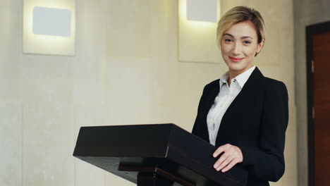 close-up view young caucasian blonde businesswoman in formal clothes on a podium looking at the camera and smiling in a conference room