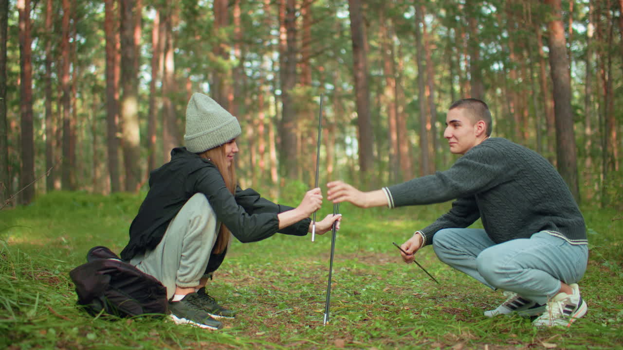 Lady playfully interacts with her man while handing over tent pole as he collects and fixes it for camping in forest, surrounded by outdoor gear and natural scenery