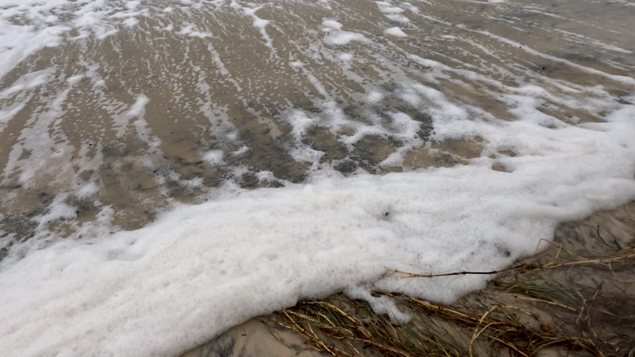 Waves crash onto a beach covered in foam and debris, highlighting environmental pollution. The scene is dynamic and unsettling