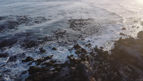 Aerial-overhead-shot-of-waves-breaking-on-rocky-seaside-shore-at-sunset,-slowly-tracking-right