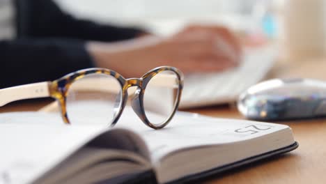 Close-up-of-personal-organizer-and-spectacle-while-businesswoman-working-over-computer