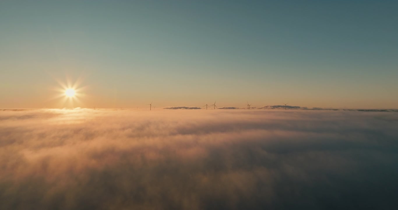 Vista aerea sopra una vasta distesa di nuvole con un bellissimo sole dorato che brilla attraverso un cielo azzurro limpido, mentre un lontano parco eolico appare all'orizzonte