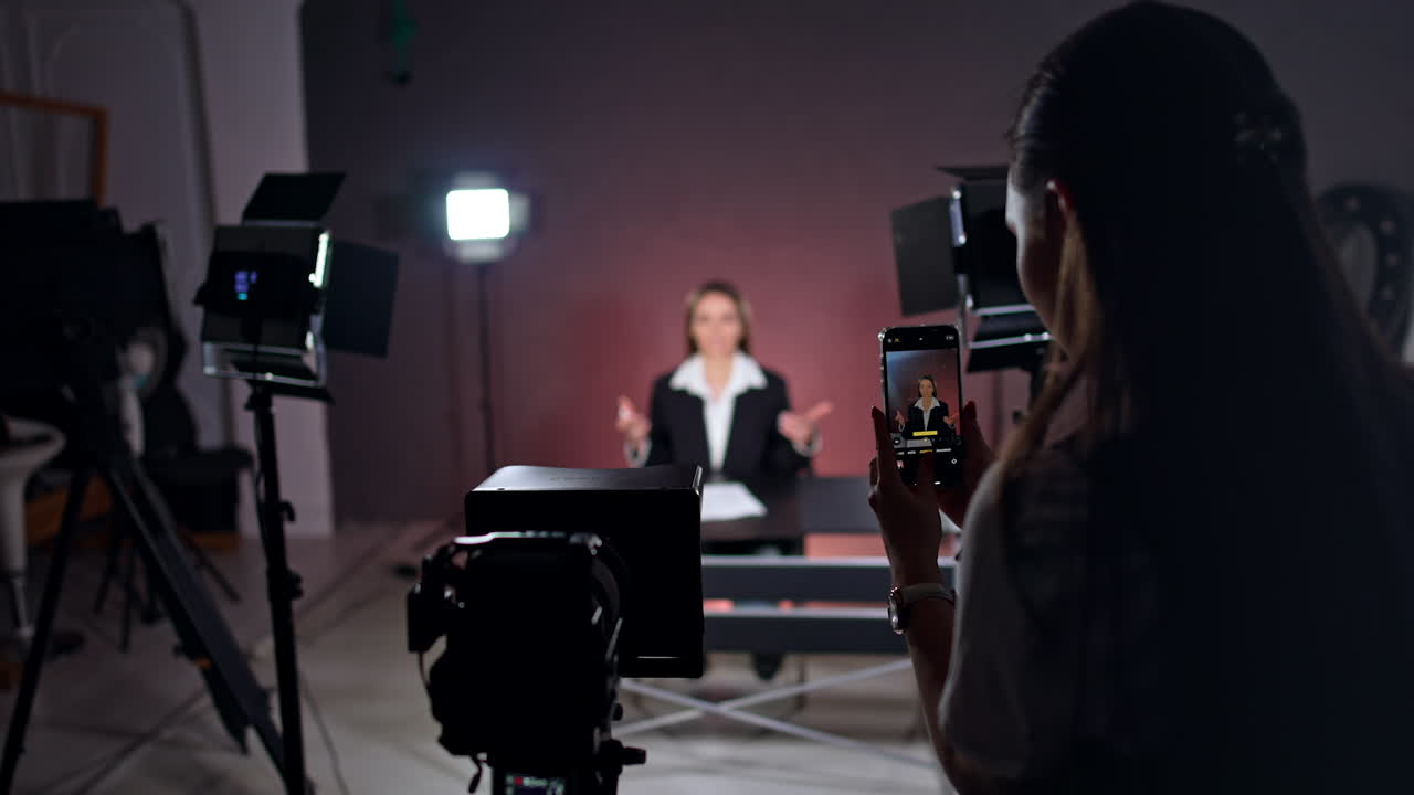 Phone footage of a lady sitting at desk in studio. Blogger is recording a video in the light of soffits. Blurred backdrop.