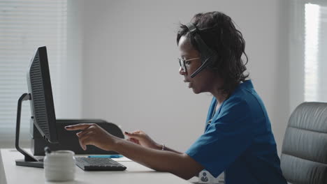 african american black female general practitioner in white coat sitting at desk in doctor's office and scrolling computer mouse while reading patient's medical history