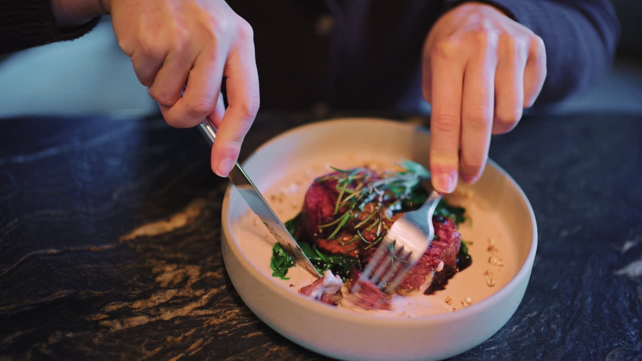 Close up of a woman's hands cutting and eating a plated steak with fork and knife