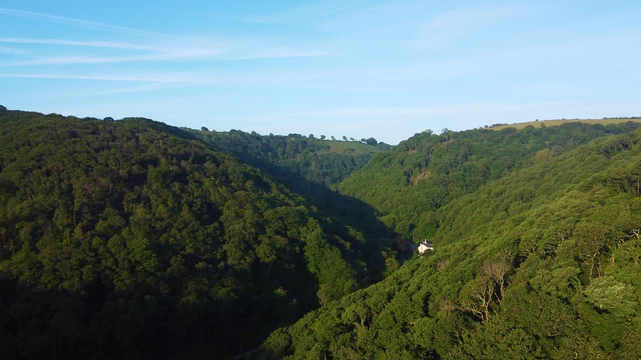 Pushing Aerial Shot of Crossing Valleys with Lone White Country House and Dense Forests Lining Hillside. Sunrise Drone Footage. Filmed in North Devon, England, UK. British Countryside.