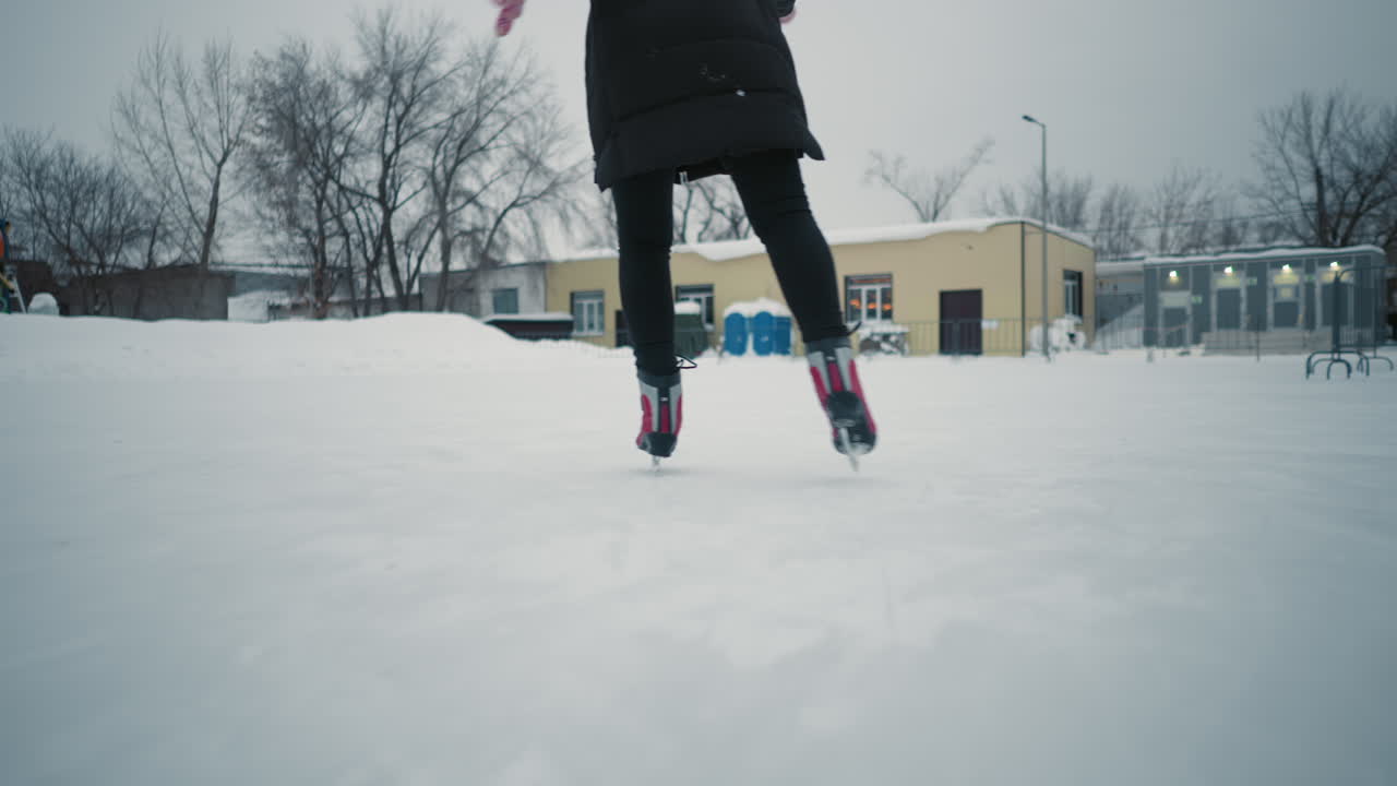 Lady gliding on outdoor ice rink in winter clothing, captured from behind focusing on red skates, black pants and mittens, with snow-covered surroundings and buildings in soft background during cold day
