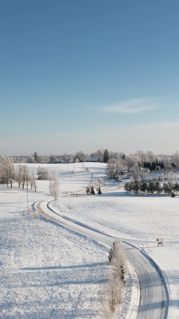Vertical aerial view of winding snow road in countryside landscape. Early morning sunrise with hoar frost on the trees. Rural area with family houses in winter season.