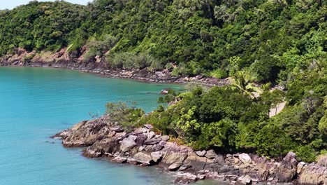 aerial drone smoothly rises above a rocky, forested coastline in port douglas, revealing turquoise water, dense rainforest, and a secluded tropical beach in bright daylight