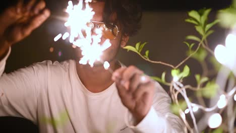 cheerful teenager male celebrates christmas, diwali or new year by playing with fireworks and crackers near led light decorated plant, slow motion