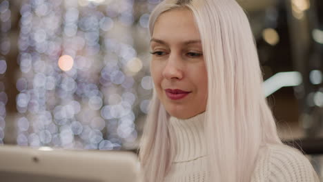 house wife seated on bench in bright mall lounge by glass railing under soft bokeh lights, focused on touchscreen tablet, smiling as she scrolls through content in cozy sweater