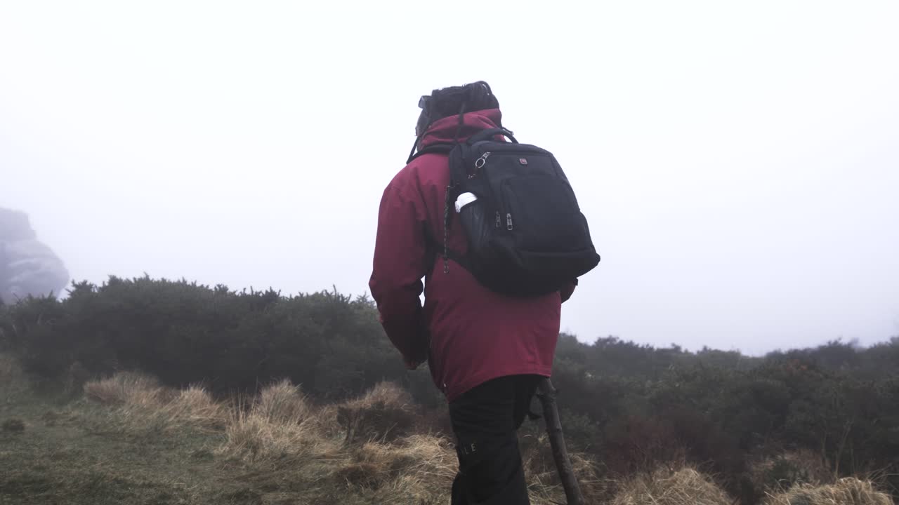 Man with a backpack and walking stick hiking towards large, weather-beaten rock formations on a foggy Dublin Mountain. Focus on wild environment, extreme weather, and solitary adventure