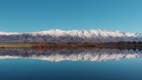 Bandada-De-Pájaros-Blancos-Volando-Sobre-Un-Lago-Tranquilo,-Con-Un-Reflejo-Perfecto-De-La-Cordillera-Y-El-Cielo-Azul