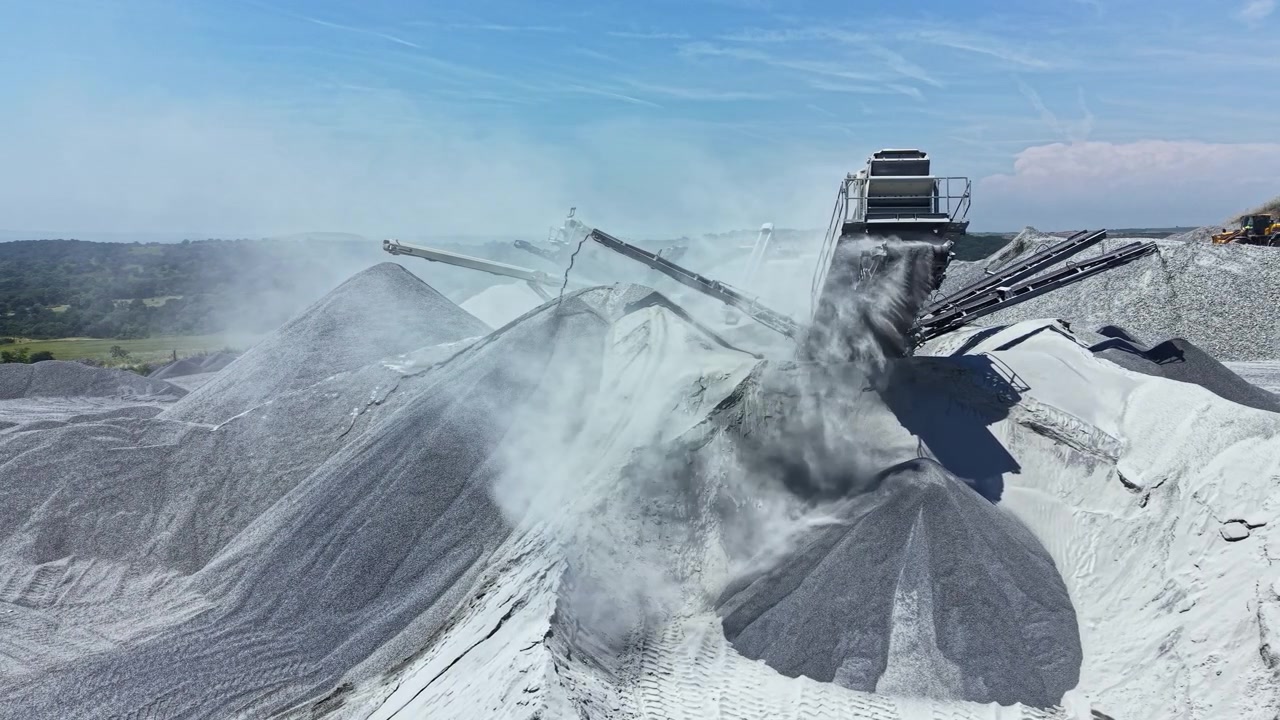 Granite quarry process showing equipment working on rock piles in open space with blue sky and dust in the air
