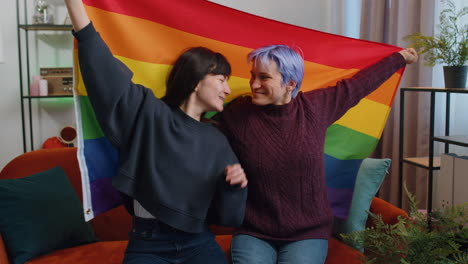 Two-lesbian-women-family-couple-or-girl-friends-holding-LGBT-people-gay-pride-flag-in-hands-at-home