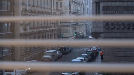 view through window blind: snow falls on narrow old world city street