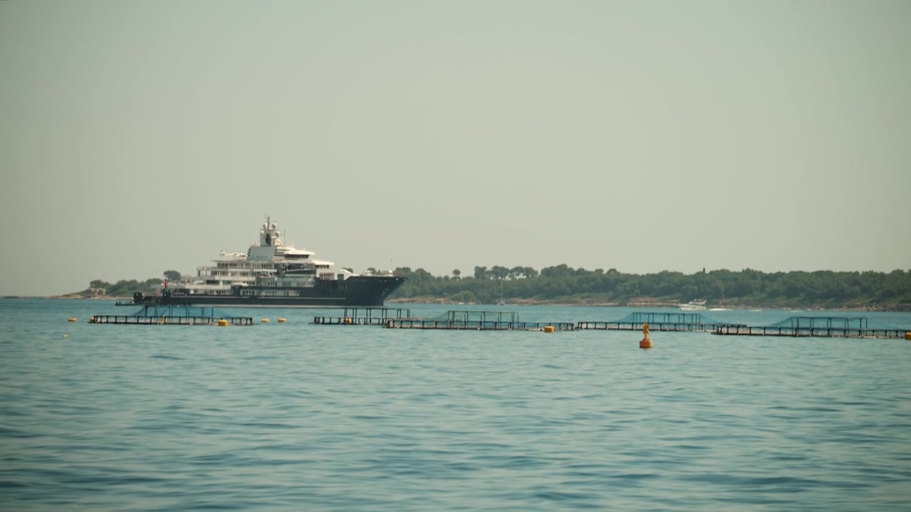 Superyacht Octopus ancorato in mare aperto con boe che galleggiano in primo piano e la costa della Costa Azzurra sullo sfondo.