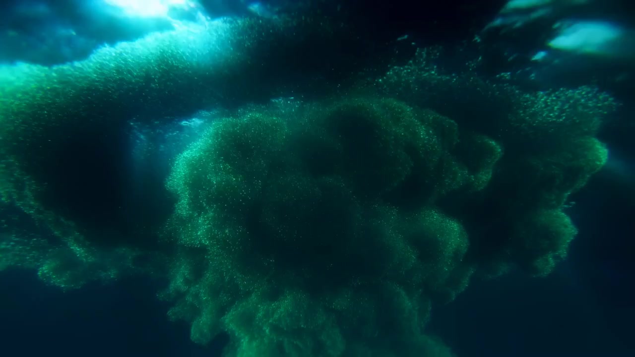 Glowing teal and green ink expands through deep dark water, creating soft swirling forms with moody low light and a dramatic abstract appearance