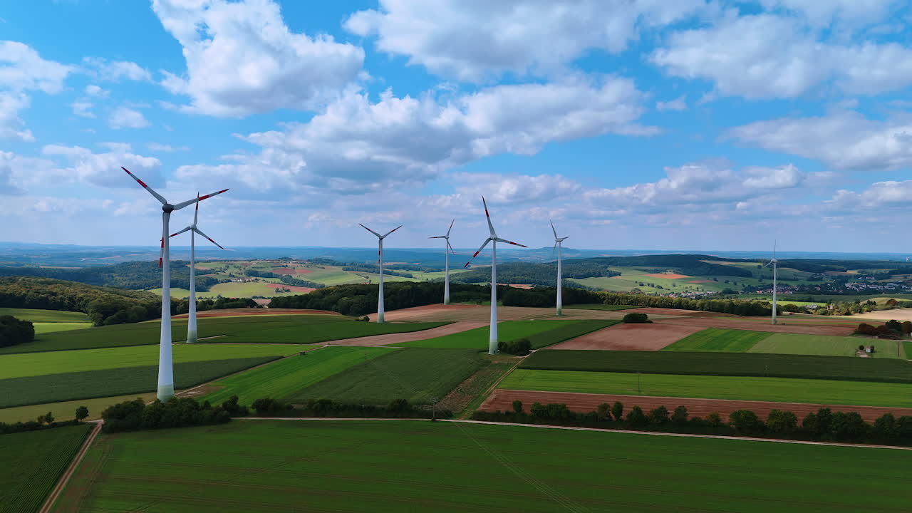 Aerial view of large wind turbines across summer countryside fields. Modern white wind turbines rising over colorful farmlands with green and brown patches under blue sky