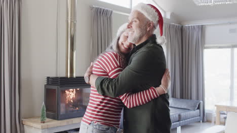 una pareja caucásica de mediana edad con sombreros de papá noel abrazándose en navidad en casa, en cámara lenta.