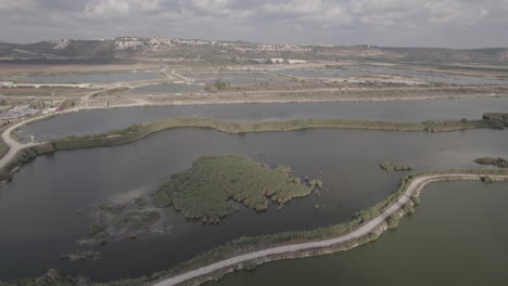 Vista-Aérea-De-Paralaje-De-Una-Isla-Con-Vegetación-En-Estanques-De-Peces-Al-Borde-De-Un-Lago-Natural-Al-Pie-De-Las-Montañas,-Una-Vasta-Y-Verde-Reserva-Natural