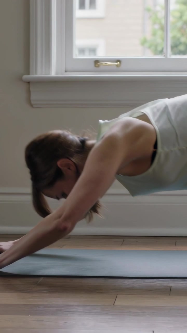 Video verticale: Donna in leggings e canotta che da una posizione piegata, spingendo le mani, si solleva in posizione plank su un tappetino vicino alla finestra.